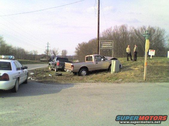 Gold pickup truck with crumpled front end, parked on grass next to a utility box. A police car is on the left