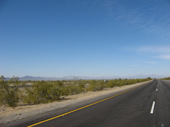 Driving on a highway through a desert landscape with scrub brush and mountains in the distance. A few cars are visible on the side of the road