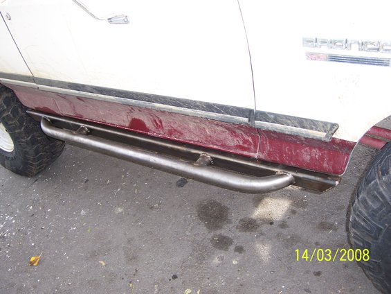Side view of a white and maroon vehicle with custom metal rock sliders installed below the doors. The word "Bronco" is visible on the body