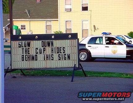 Police car parked next to a sign that reads 'SLOW DOWN THE COP HIDES BEHIND THIS SIGN'