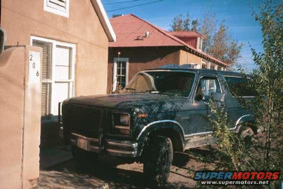 Dark Ford Bronco parked next to a tan house. Trees and bushes are in the foreground and to the right