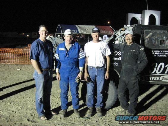 Four men in casual and racing attire stand next to a 1994 Ford Bronco race truck with "301" and "RIVER SUSPENSION" visible