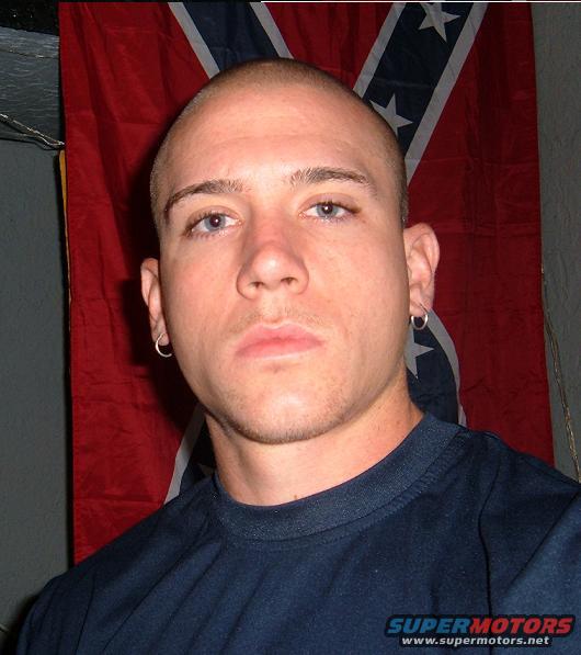man in navy shirt, hoop earrings, bald head, in front of confederate flag. Supermotors.net logo in lower right