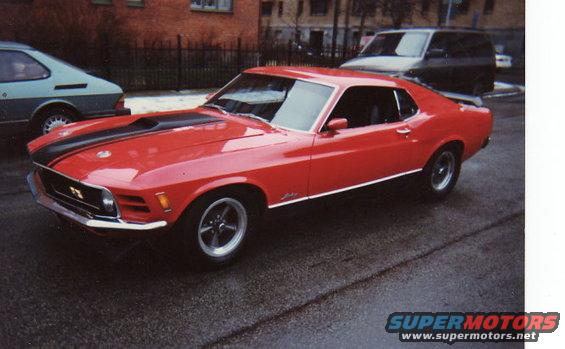 Red 1970 Ford Mustang coupe with black hood stripes and chrome trim, parked on wet asphalt