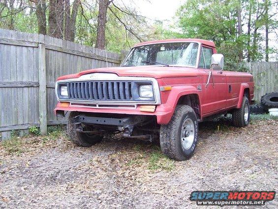 Red pickup truck parked in front of a wooden fence. Front bumper is missing