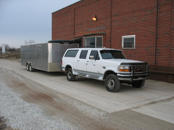 White Ford F-250 extended cab truck towing a silver enclosed trailer, parked next to a brick building