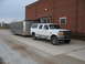 1995 Ford C-350 (Centurion Conversions) - White Ford truck with a silver trailer is parked on a gravel and concrete surface near a brick building.