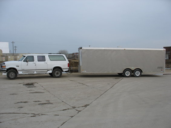 White Ford Bronco SUV towing a silver enclosed trailer. The SUV has four doors and a custom rear section