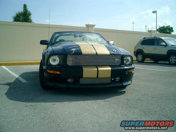Black convertible Mustang with gold stripes parked in a lot, another SUV is behind it