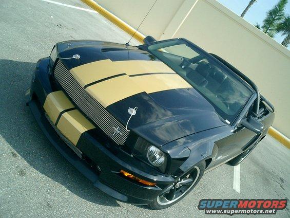 Black convertible with gold racing stripes parked on asphalt. The car has a black grille and gold accents on the hood