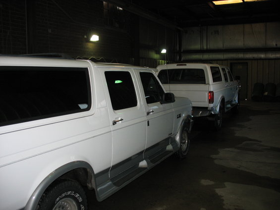 Two white trucks parked in a garage. The one in front is an extended cab pickup, and the one behind it is a crew cab pickup with a camper shell