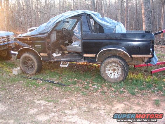 Black Ford Bronco with driver door open, covered partially with a tarp. Another vehicle is partially visible on the left