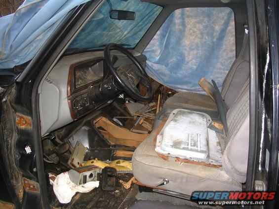 Interior of a black 1980s Ford Bronco with torn upholstery and debris scattered on the floor, covered by a blue tarp