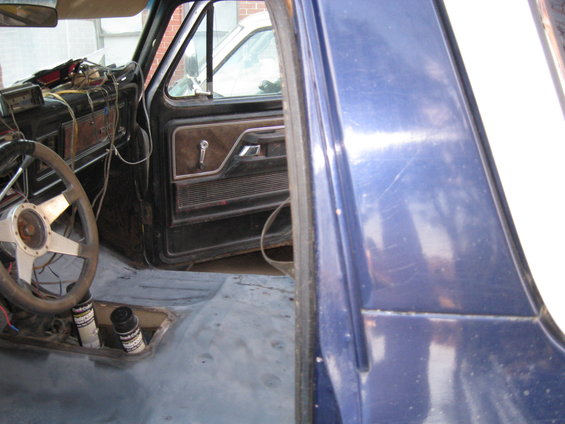 Interior of a truck with exposed floor, aftermarket steering wheel, and door panel visible. Another vehicle is seen through the window