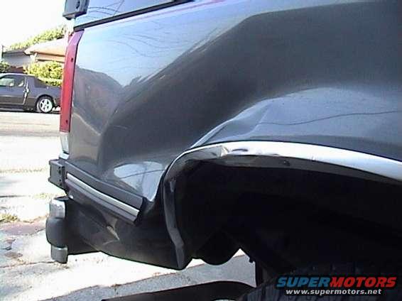 Gray truck rear quarter panel with significant damage. Another car is visible in the background