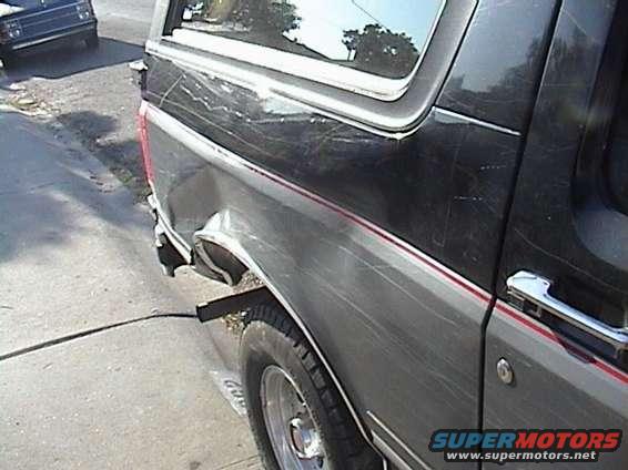 Rear driver's side of a dark-colored SUV with a dented quarter panel and a red stripe. Another vehicle is visible in the background