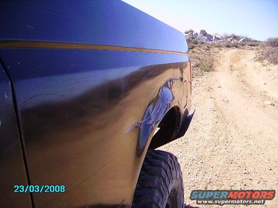 Rear quarter panel of a dark vehicle with significant damage, showing a dirt road and rocky terrain