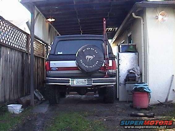 Rear view of a 1990 Ford Bronco parked under a carport. It has a spare tire on the back and a Hi-Lift jack mounted