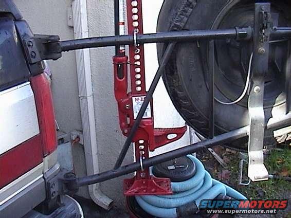 Hi-Lift jack attached to a spare tire mount on a vehicle's rear. Blue hose coiled below