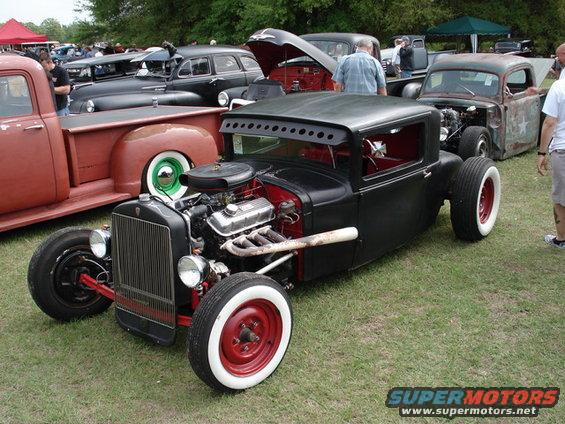 Black hot rod pickup truck with exposed engine, red wheels, and white wall tires, viewed from the front-right