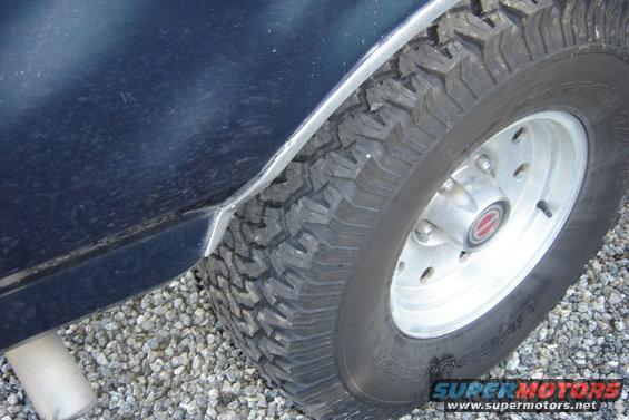 Close-up of a dark blue vehicle's rear tire and fender area. The tire has aggressive tread. A silver exhaust pipe is visible below