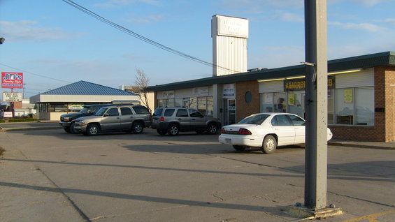 Commercial buildings and parking lot with several cars including a white sedan and SUVs