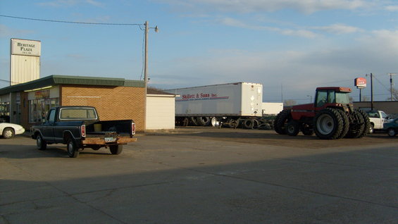Dark blue pickup truck parked in front of a building, with a semi-trailer and a red tractor behind it