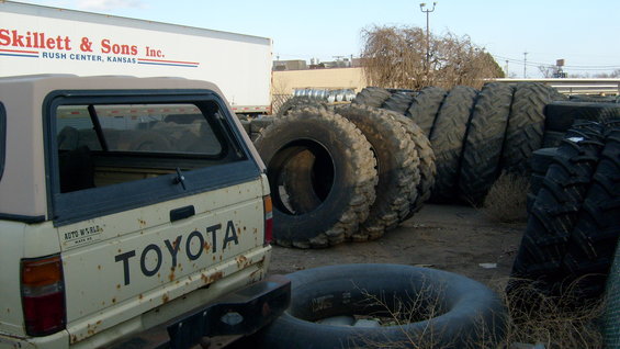 Rear view of a Toyota pickup truck with a camper shell, next to large tires and a semi-trailer