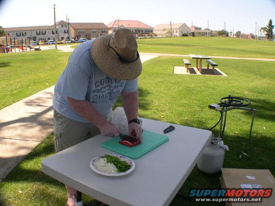 Man wearing a straw hat chops red bell pepper on a cutting board at a picnic table in a park