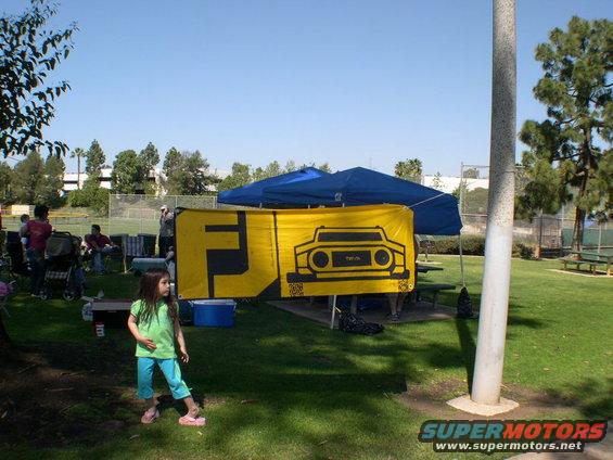 Yellow banner with FJ Cruiser graphic and text, at an outdoor gathering with people and tents