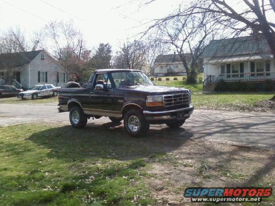 Dark-colored Ford Bronco parked on a dirt driveway in front of houses
