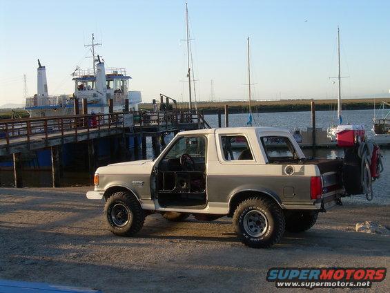 1991 Ford Bronco parked by a dock with boats and a ferry in the background