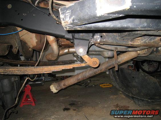 Close-up of vehicle undercarriage showing leaf springs, exhaust pipe, and suspension parts. A red jack stand is visible