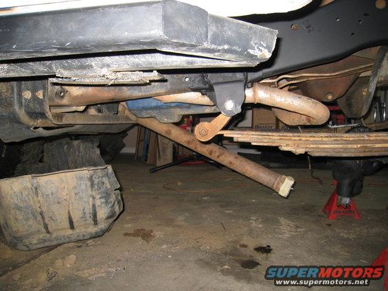 Underside of a vehicle showing suspension components, leaf springs, and exhaust pipe. A fuel tank is visible on the left