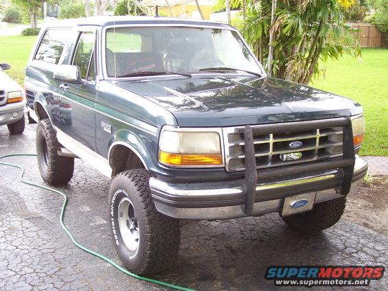 Green Ford Bronco with a brush guard and aftermarket wheels, parked on a driveway. Another vehicle is partially visible to the left