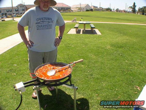 Man in a hat and t-shirt stands by a large pan of food on a burner in a grassy park