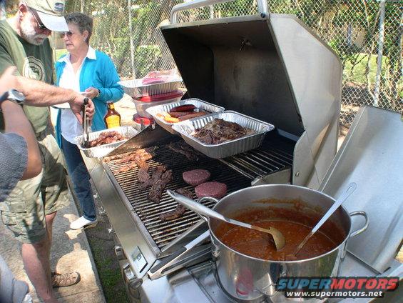People are grilling burgers, ribs, and hot dogs on a large gas grill. A pot of chili sits to the side