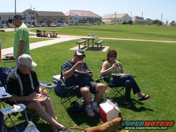 People enjoying a picnic in a grassy park with picnic tables and buildings in the background