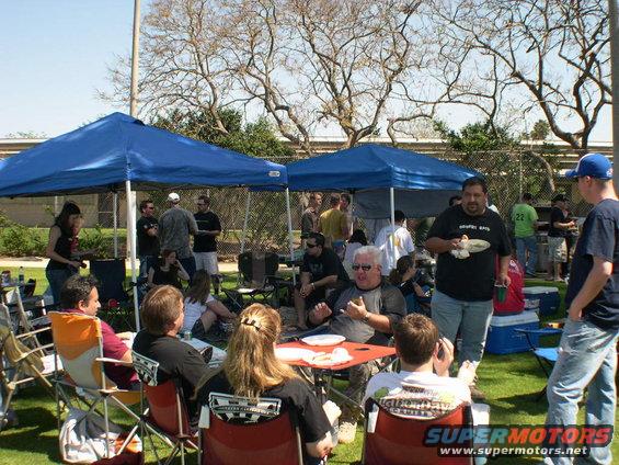 People gathered outside under blue tents, some sitting in chairs, others standing and talking. Food and drinks are visible