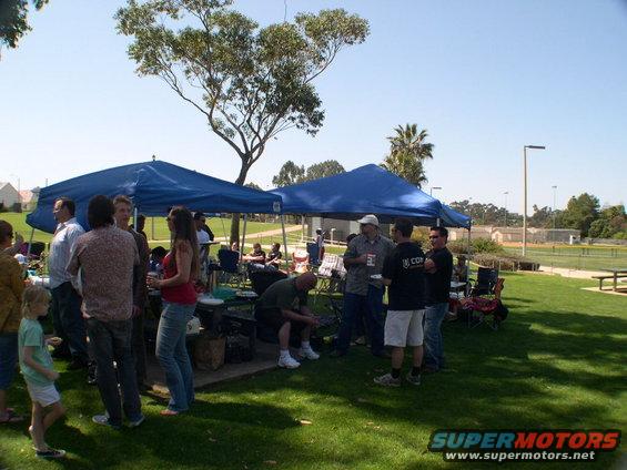 People gathered under blue tents on a sunny day at a park with a chain-link fence and palm trees in the background
