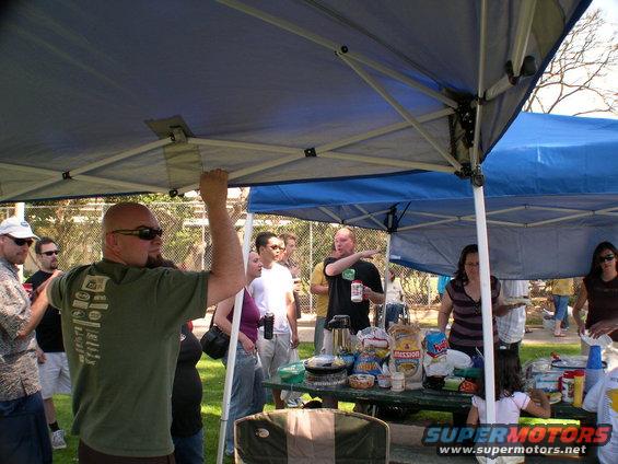 People gathered under blue and green tents at an outdoor event with a picnic table laden with food