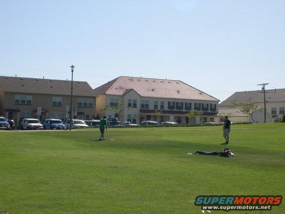 Several cars parked in a lot behind a grassy field with buildings in the background