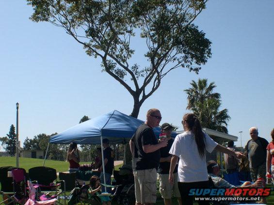 People gathered under a canopy at an outdoor event with trees and a grassy field