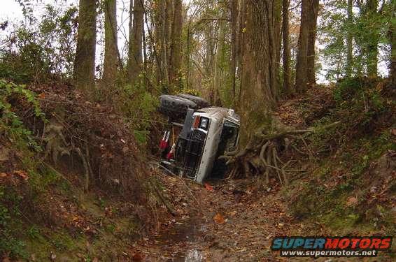 White SUV overturned on its side in a wooded ditch, front end visible