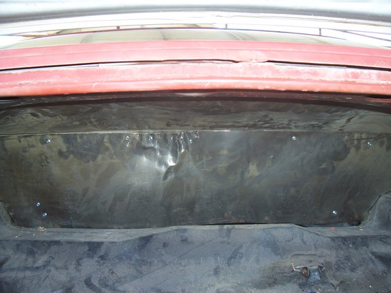 Car trunk interior showing a dented metal panel screwed into the back wall, with black carpet lining the floor