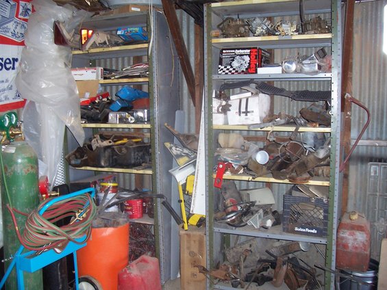 shelving units packed with car parts, including carburetors, engine components, and miscellaneous items. A welding tank and gas cans are also visible