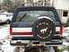 1988 Ford Bronco - Rear view of a 1988 Ford Bronco in a snowy setting, showing the spare tire cover and a white car in the background.