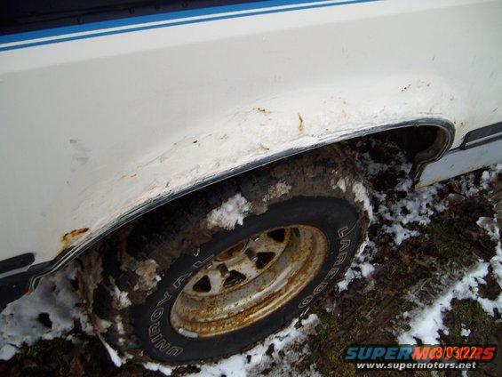 Close-up of rusty wheel well and tire on a white vehicle with a blue stripe, in snowy, muddy conditions