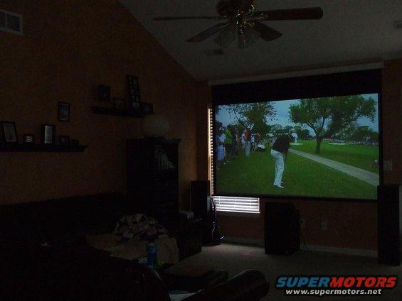 Projector screen shows a golf game with a player swinging. Room has a couch and fan