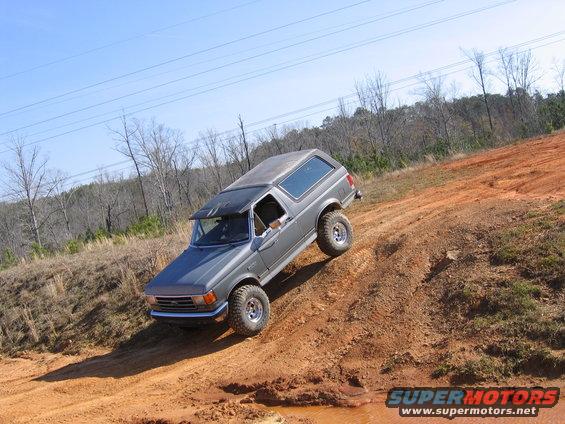 Gray SUV with aftermarket wheels driving up a muddy dirt hill, clear blue sky